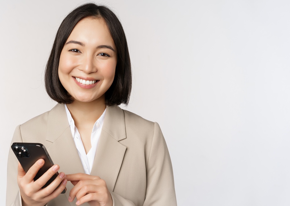 Woman in a beige blazer holding a smartphone and smiling, standing against a plain white background.