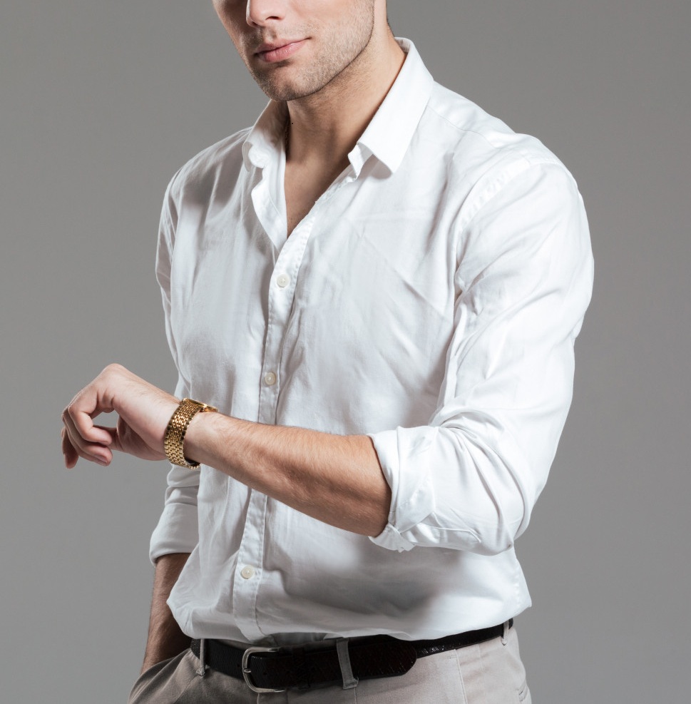Man wearing a white button-up shirt and beige pants looks at a gold wristwatch, with his left arm slightly raised. The background is plain and gray.