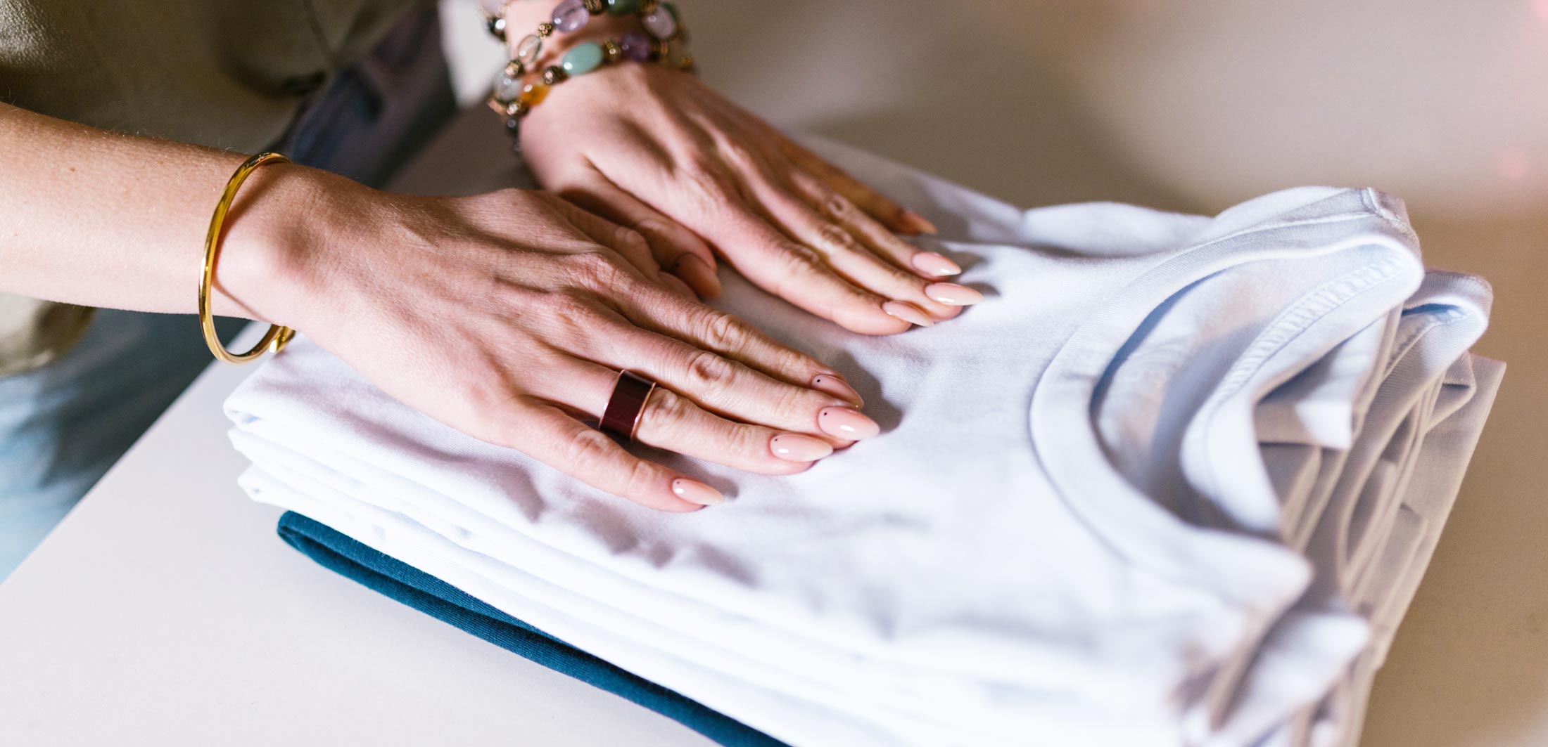 A person with manicured nails folds a stack of neatly arranged t-shirts on a white surface.