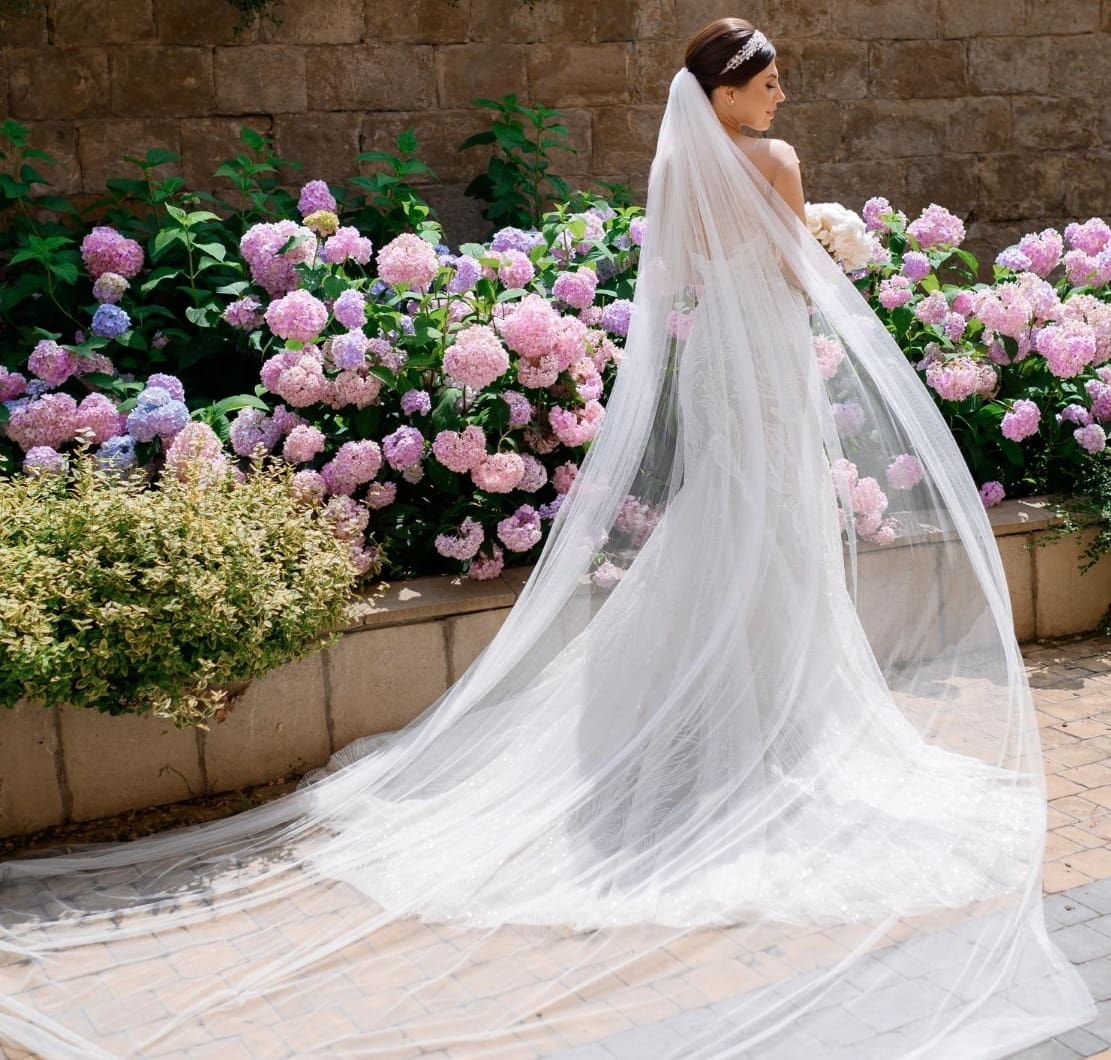 Bride in elegant wedding gown with flowing veil, surrounded by colorful hydrangeas in a garden setting, representing Craft Cleaners' wedding gown cleaning and preservation services.