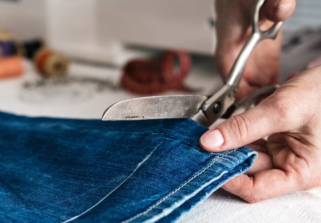 Tailor cutting denim jeans to shorten length while preserving original hem details, with sewing tools visible in the background.