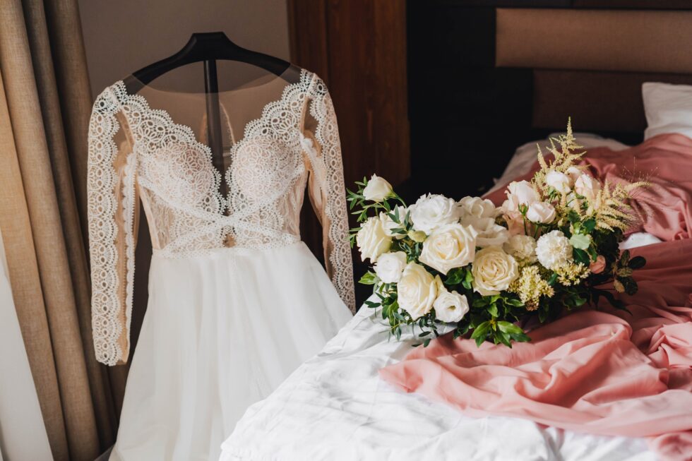 Wedding dress with intricate lace detailing hanging on a hanger beside a bouquet of white and pastel flowers on a bed with pink fabric, symbolizing preparation for the big day and clothing care.