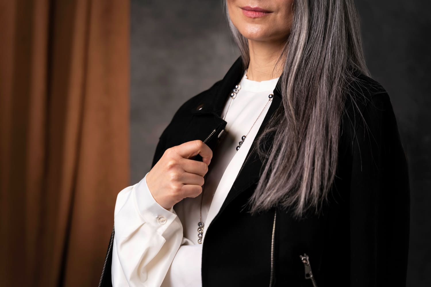 A person with long gray hair wears a black zip-up jacket over a white shirt, holding the jacket collar with one hand. The face is partially out of frame.