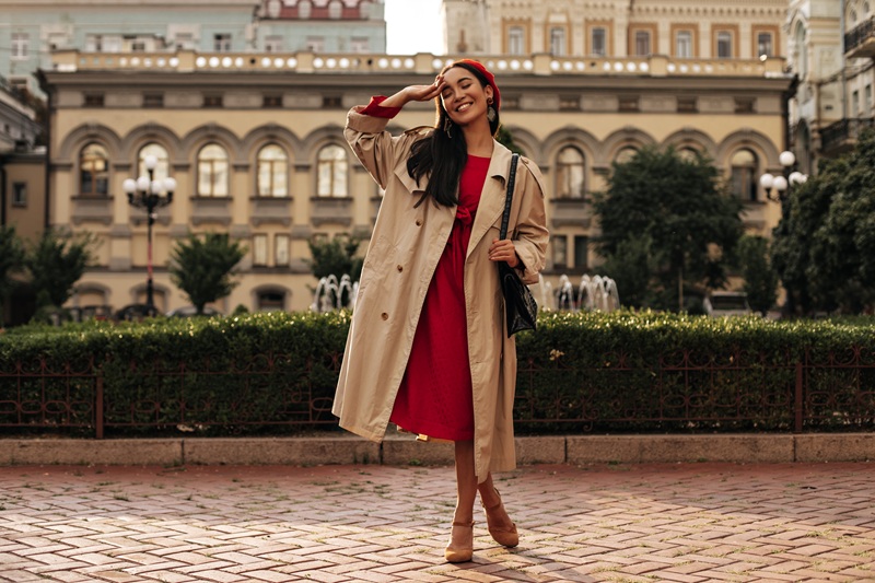 Woman wearing a red dress, beige trench coat, and red beret poses and smiles in front of a historic building and fountain in an urban setting.