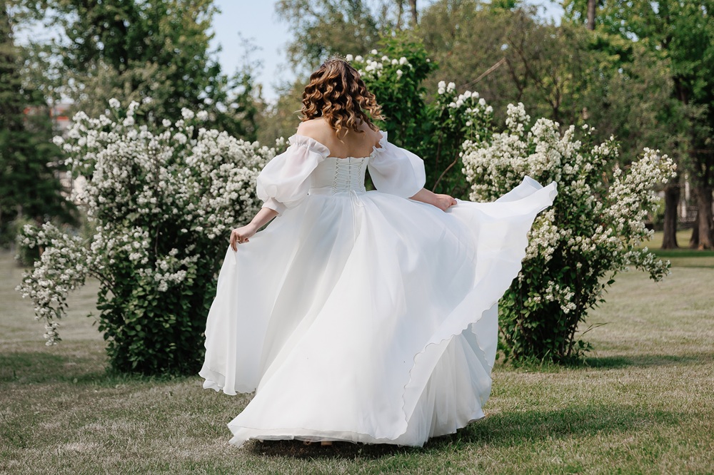 A woman in a white off-the-shoulder gown stands outdoors on grass, facing away, surrounded by blooming bushes and trees.