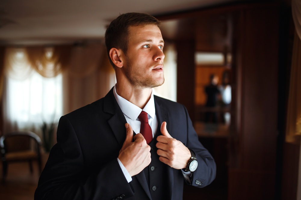 A man in a dark suit and red tie stands indoors, adjusting his jacket and looking to the side.
