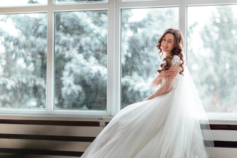 A woman in a white wedding dress sits by large windows with a snowy landscape outside, smiling slightly and looking at the camera.
